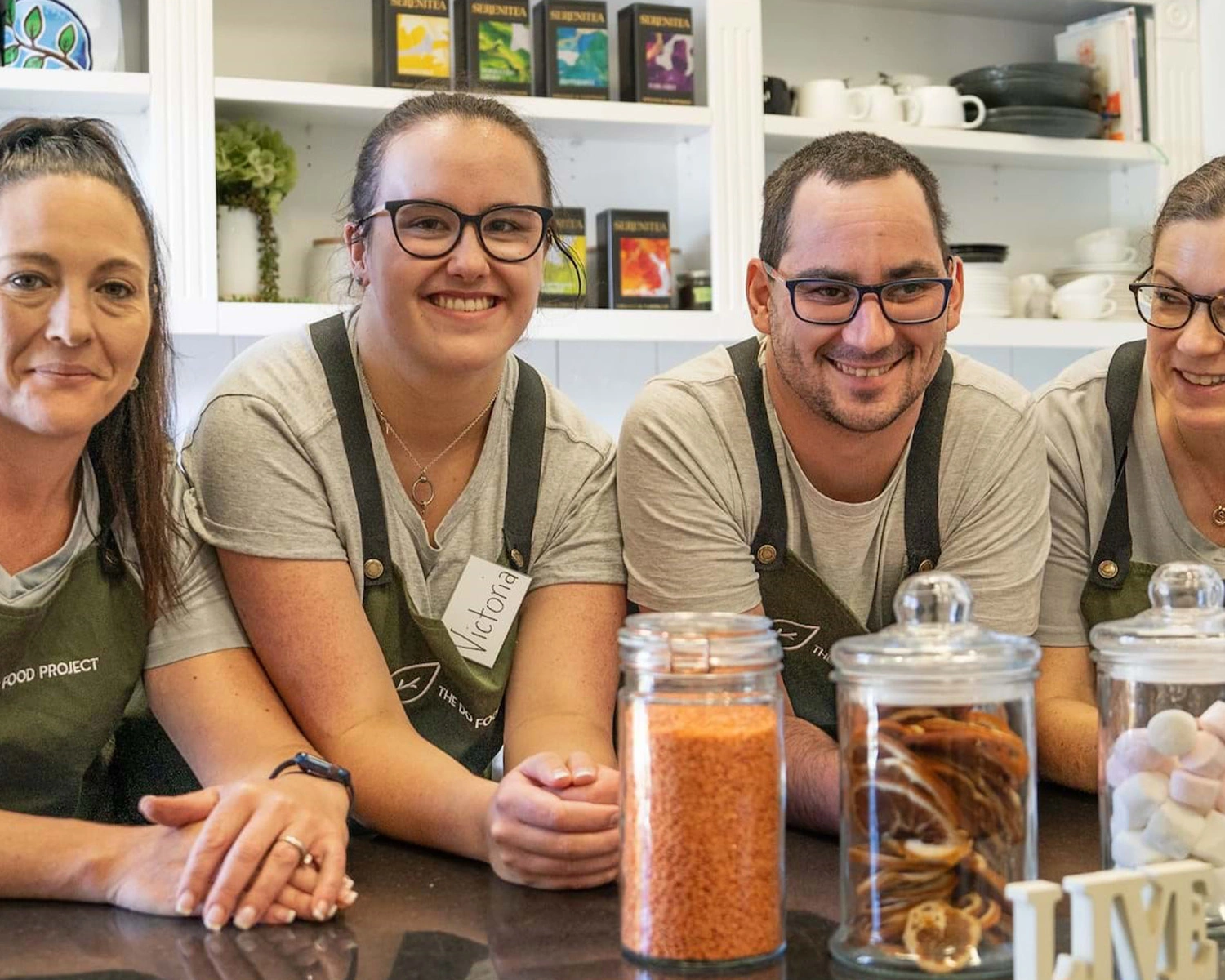 Four people wearing cafe aprons smile while leaning on a counter at NDIS registered disability service Distinctive Options