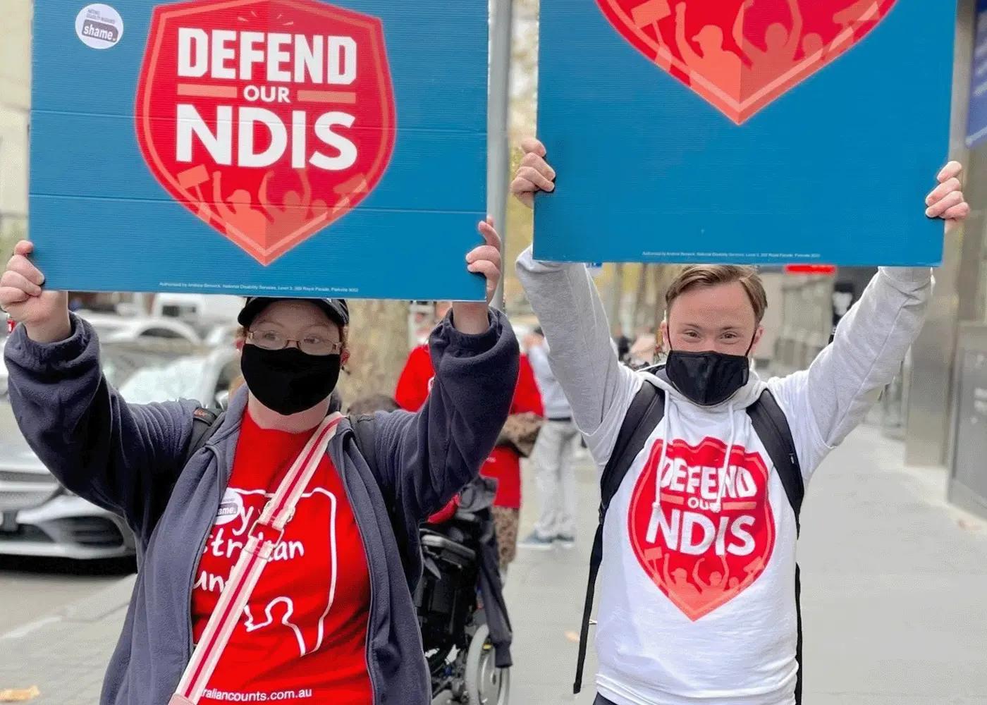 Two young people stand on the street holding up signs saying "Defend Our NDIS."